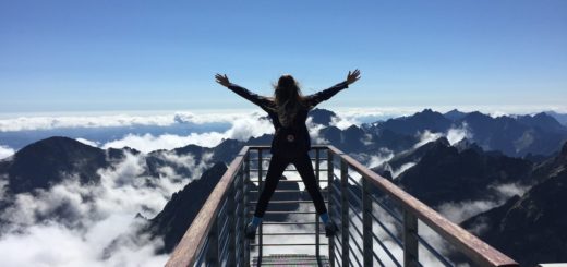 person standing on hand rails with arms wide open facing the mountains and clouds